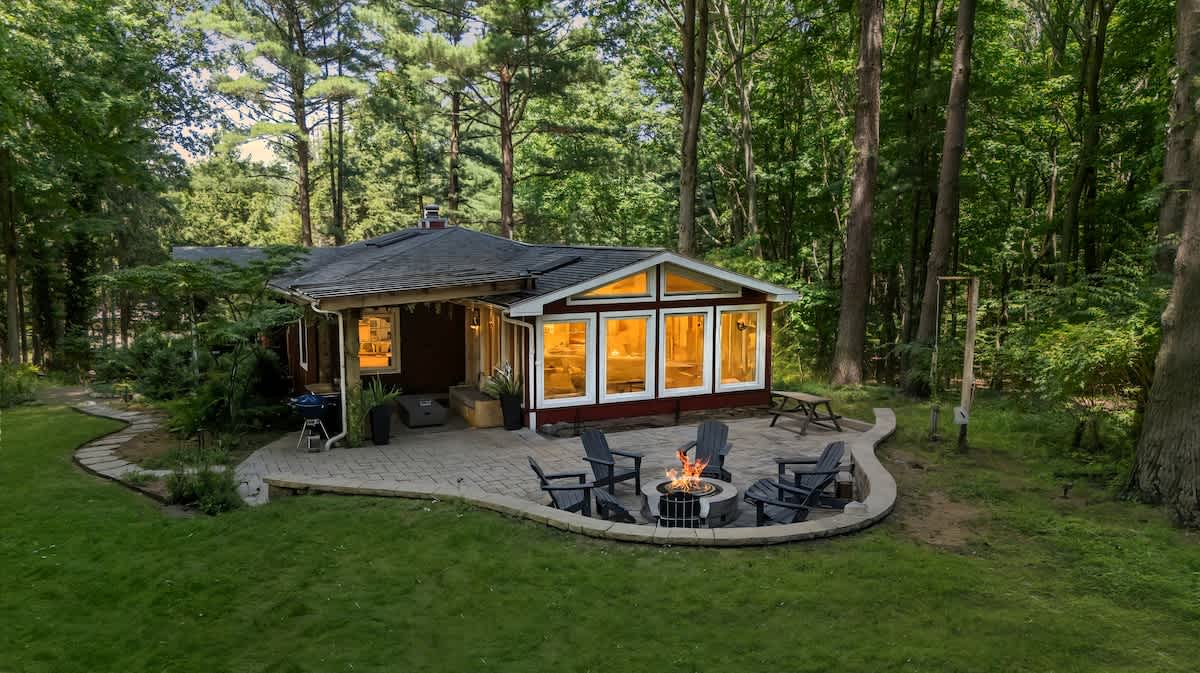 Aerial view of a charming cottage nestled among tall trees, featuring a stone patio with a fire pit and outdoor seating. The sunroom with large windows is prominent on the side.