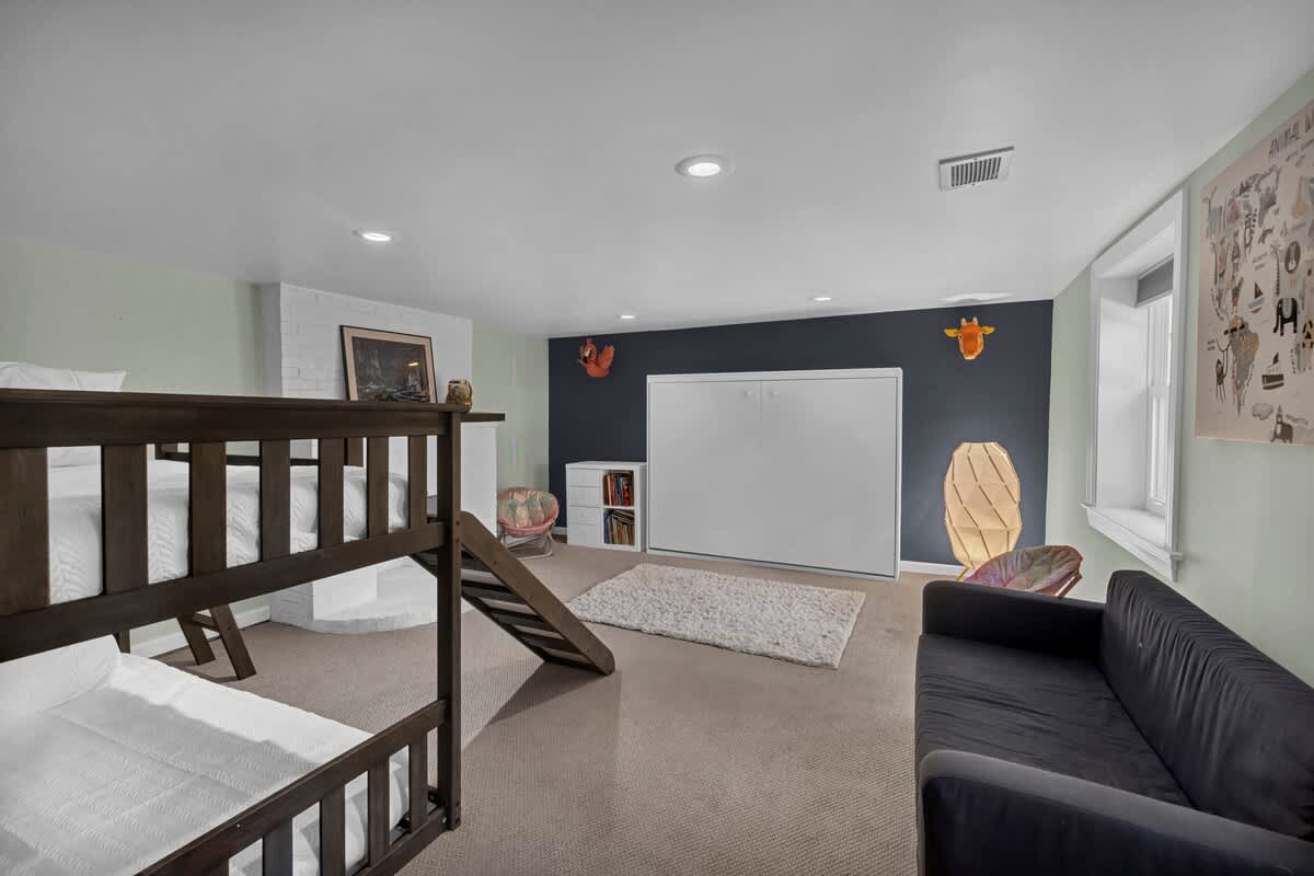A versatile bedroom with a wooden bunk bed, a dark grey sofa, and a white Murphy bed integrated into a dark blue accent wall. The room features light green walls, carpet flooring, and decorative animal head mounts.