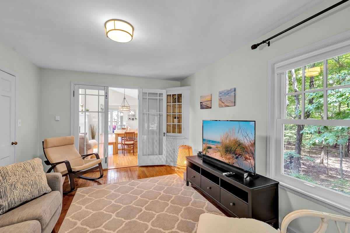 Bright living area with light blue walls, featuring a large flat-screen TV on a dark console. French doors open to another room, and a window offers views of the surrounding trees.