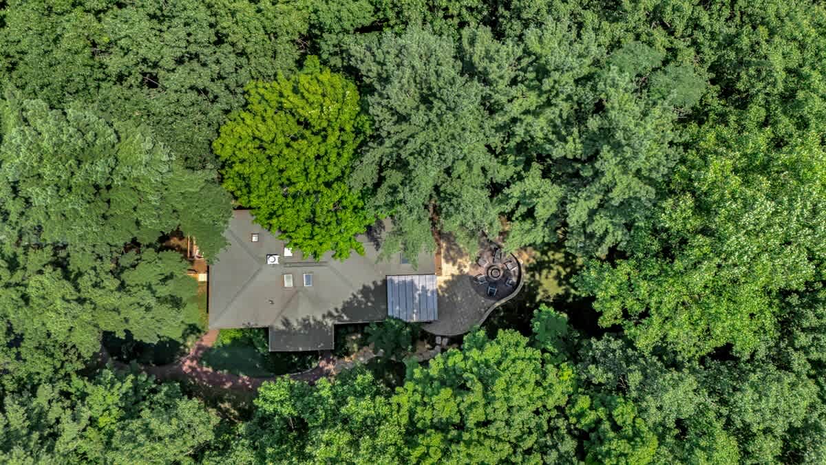 An aerial view showcasing the house nestled within a dense forest of green trees. The roof of the house and a portion of the outdoor patio with a fire pit are visible through the canopy.