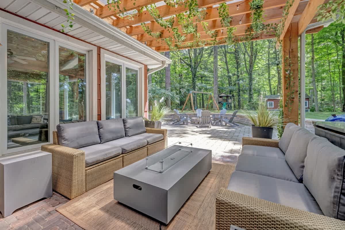Inviting outdoor patio under a wooden pergola draped with vines, featuring comfortable wicker sofas and a modern fire pit coffee table. A lush forest and a children's play area are visible in the background.