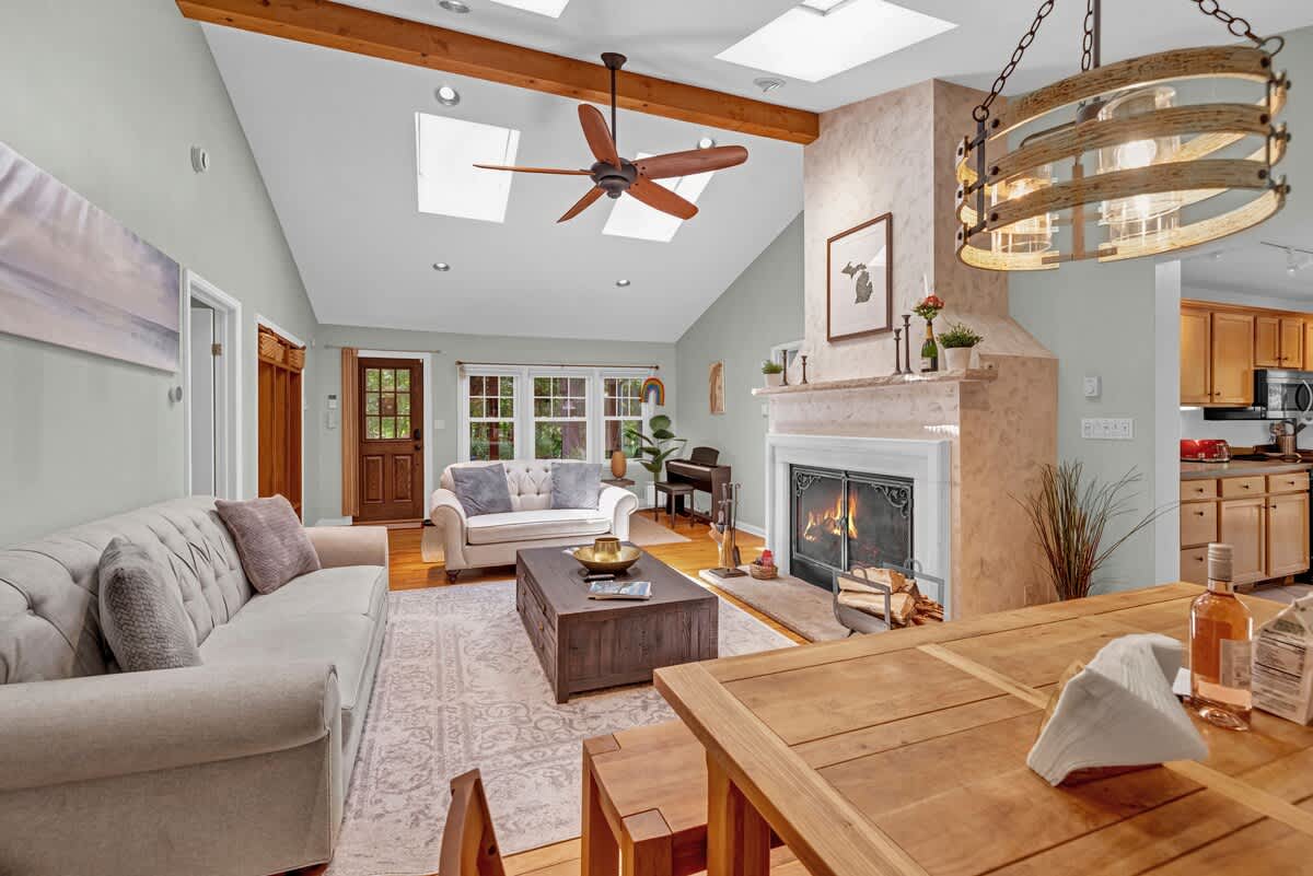 Cozy living room with a vaulted ceiling, skylights, and a prominent fireplace with a mantel. Light-colored sofas are arranged around a dark wood coffee table, with a dining area visible beyond.