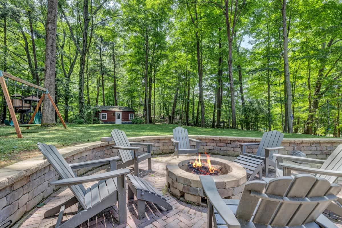 A cozy outdoor fire pit area featuring a circular stone fire pit surrounded by grey Adirondack chairs on a brick paver patio. The area is enclosed by a low stone wall and backed by lush green trees.