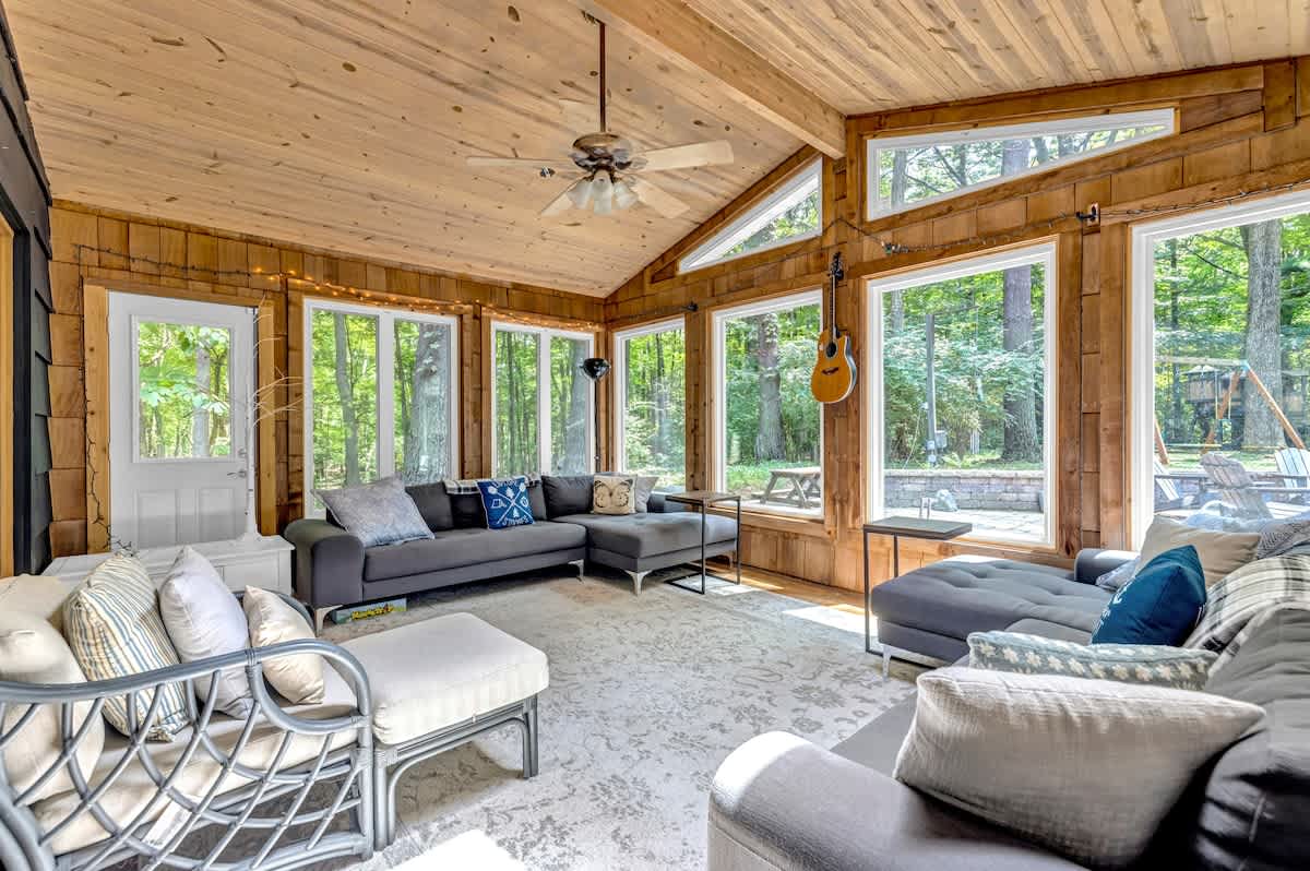 Expansive sunroom with a vaulted wooden ceiling and abundant natural light from numerous windows. Multiple gray sectional sofas and ottomans provide ample seating on a large patterned rug.