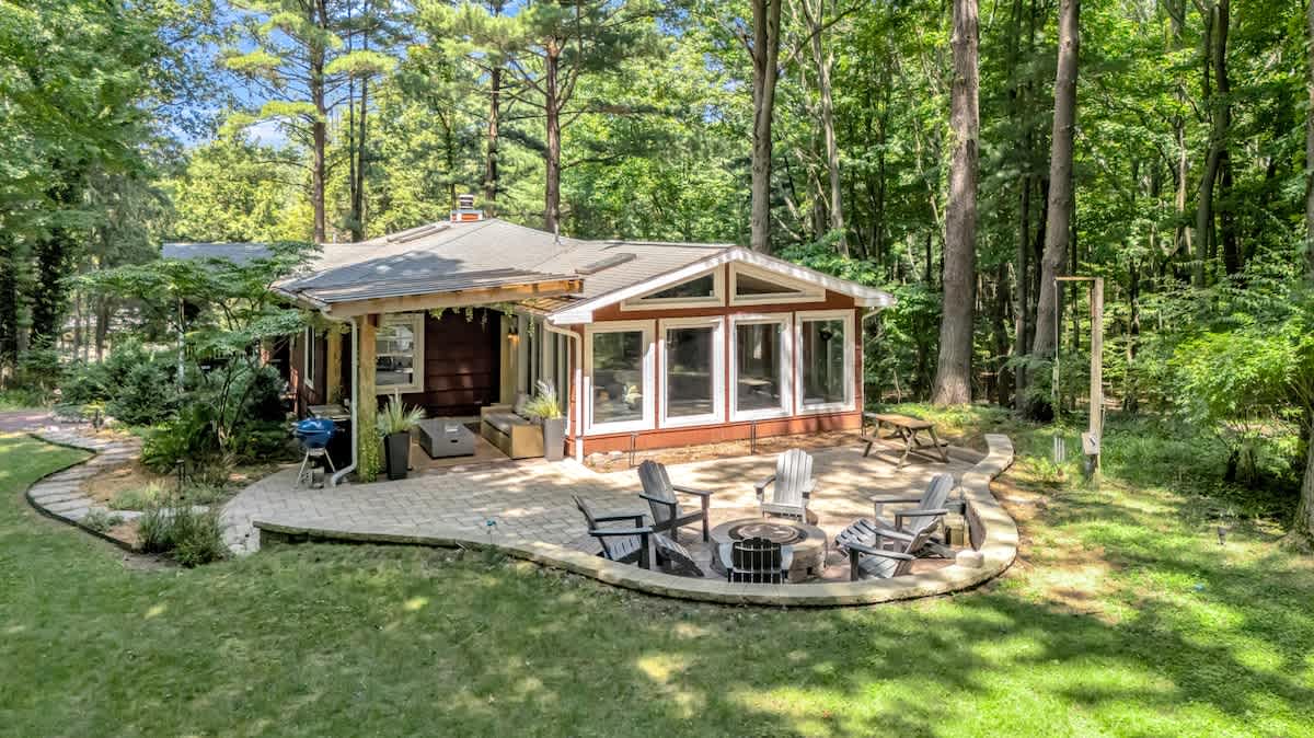 The back exterior of the house, featuring a large stone patio with a circular fire pit area surrounded by Adirondack chairs. The house has red siding and large windows, nestled among tall green trees.