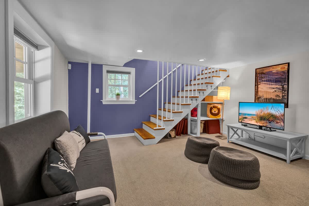 A cozy recreation area with a gray futon, a television on a white console, and stairs leading up, featuring a purple accent wall and carpeted flooring.