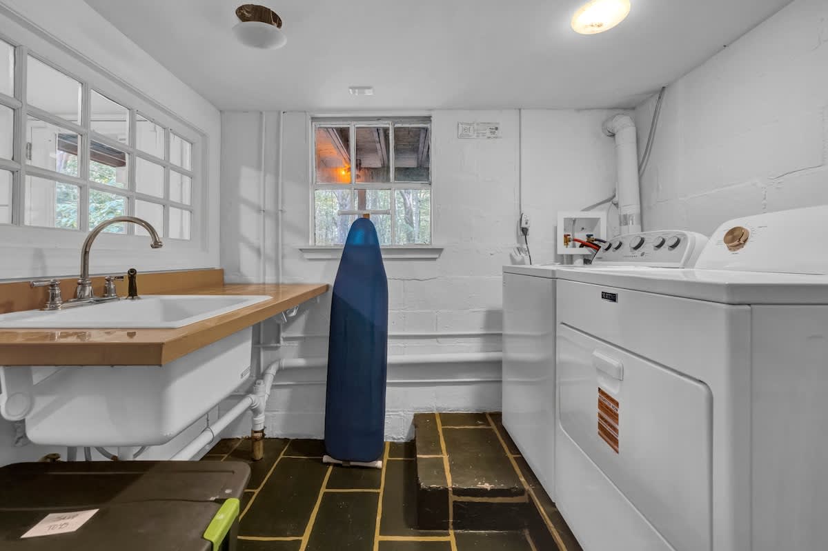 A functional laundry room equipped with a white washer and dryer, a utility sink with a wooden countertop, and dark slate-like floor tiles. The room has white painted brick walls and multiple windows.