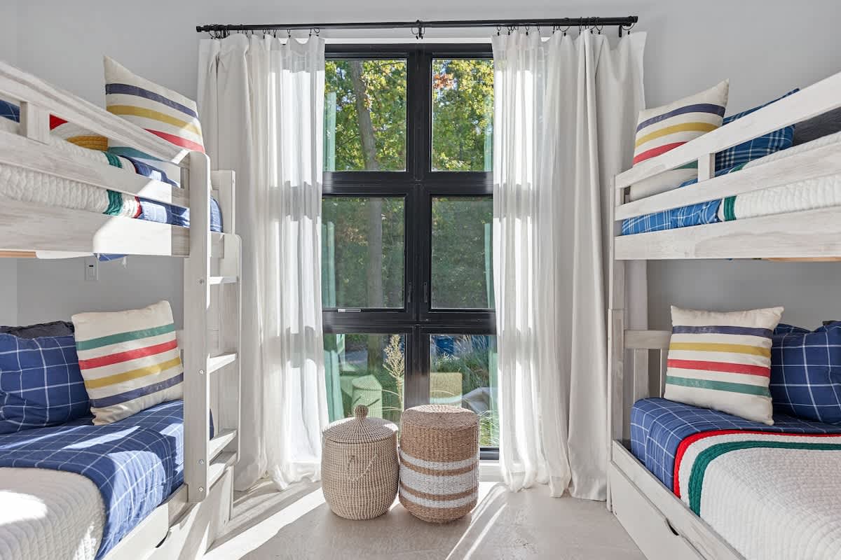 Symmetrical view of a kids' bedroom with two white wooden bunk beds, each adorned with colorful striped pillows and blue bedding, flanking a large black-framed window with white curtains and two woven storage baskets.