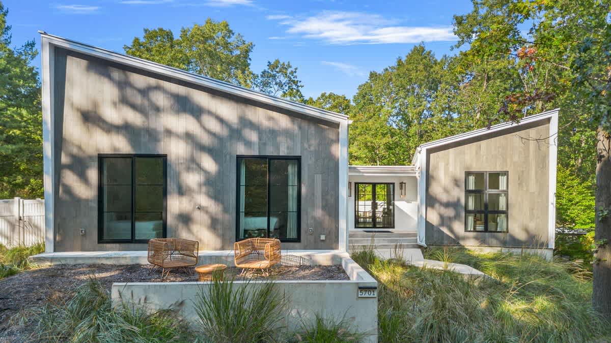 The modern front exterior of the house features light grey wood siding and large black-framed windows, with a distinctive angled roofline. Lush green landscaping and a concrete retaining wall with the house number "5701" are visible.