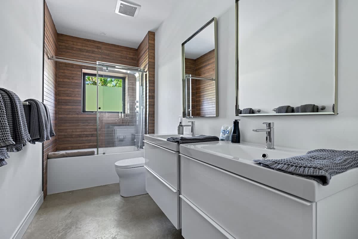 Contemporary bathroom with a double white floating vanity, rectangular mirrors, and a bathtub/shower combo with wood-paneled walls and a frosted window.