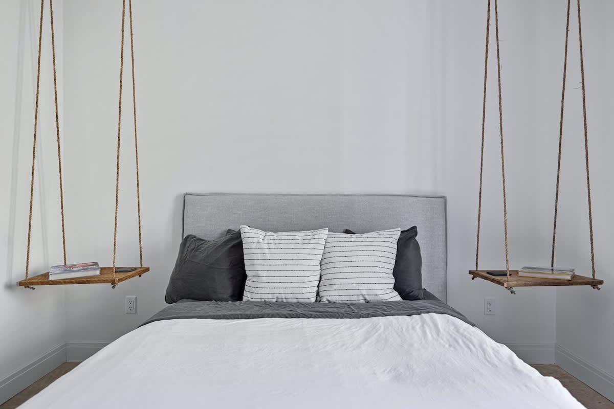 Close-up of a gray upholstered bed with white and striped pillows, flanked by two rustic hanging wooden shelf nightstands suspended by rope, against a plain white wall.