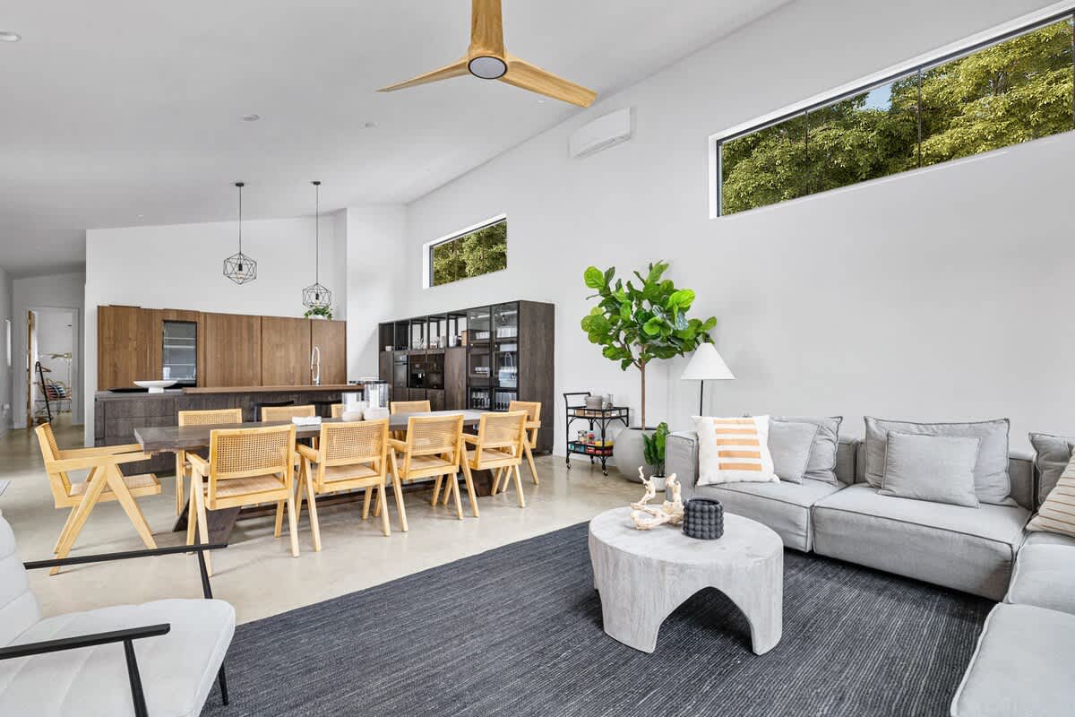 Open-concept living space featuring a gray sectional sofa, a concrete coffee table, and a large dark rug, flowing into a dining area with a wooden table and cane chairs, and a modern kitchen with dark wood cabinetry.