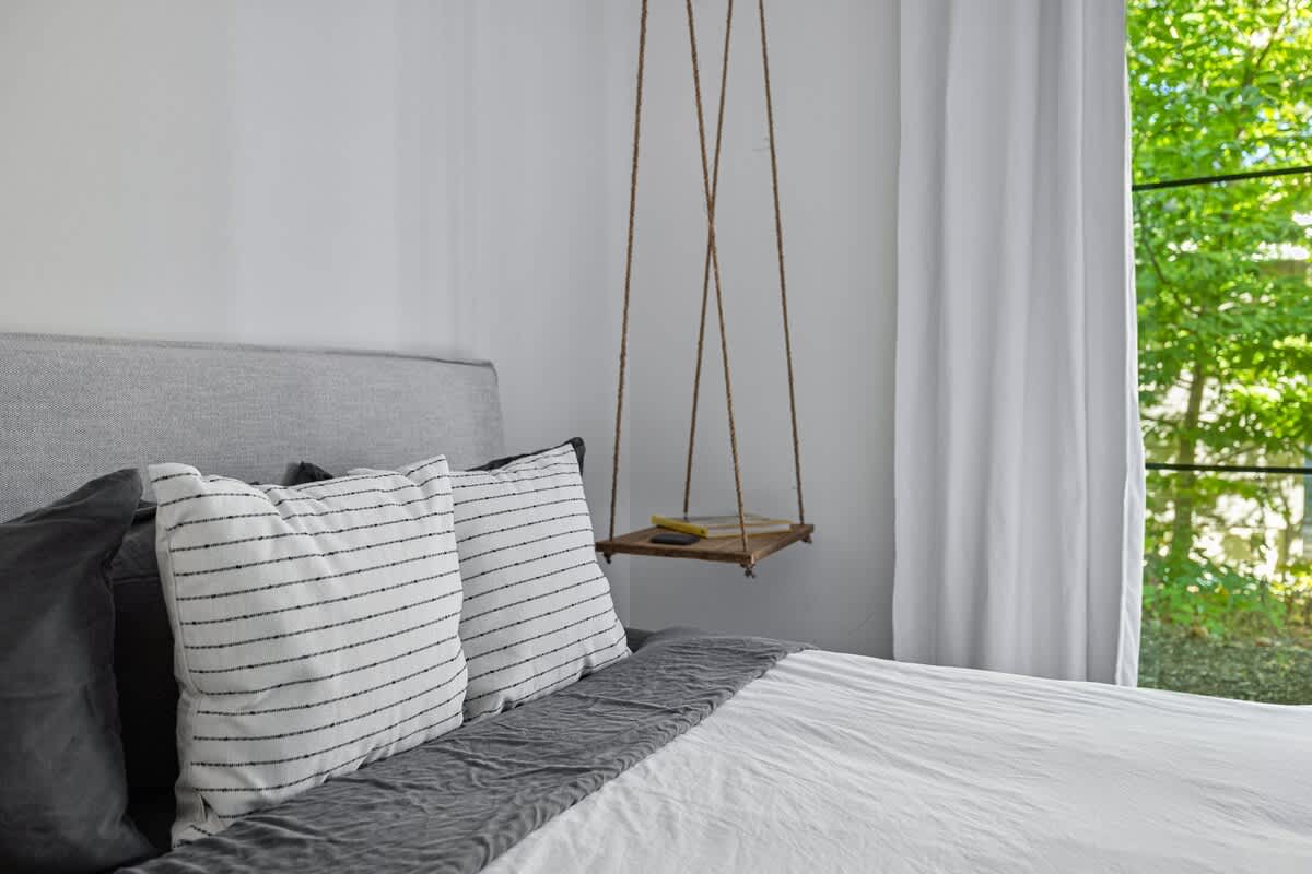 Detail shot of the bed with white bedding and striped pillows, showing one of the unique rope-suspended wooden shelf nightstands with a book, next to a large window with white curtains.
