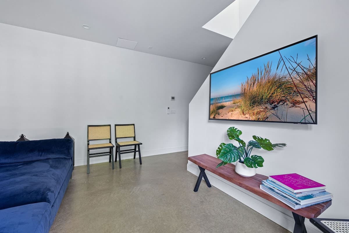 A minimalist living space featuring a large wall-mounted TV displaying a beach scene, a wooden bench with a plant and books, and two cane-back chairs against a white wall.