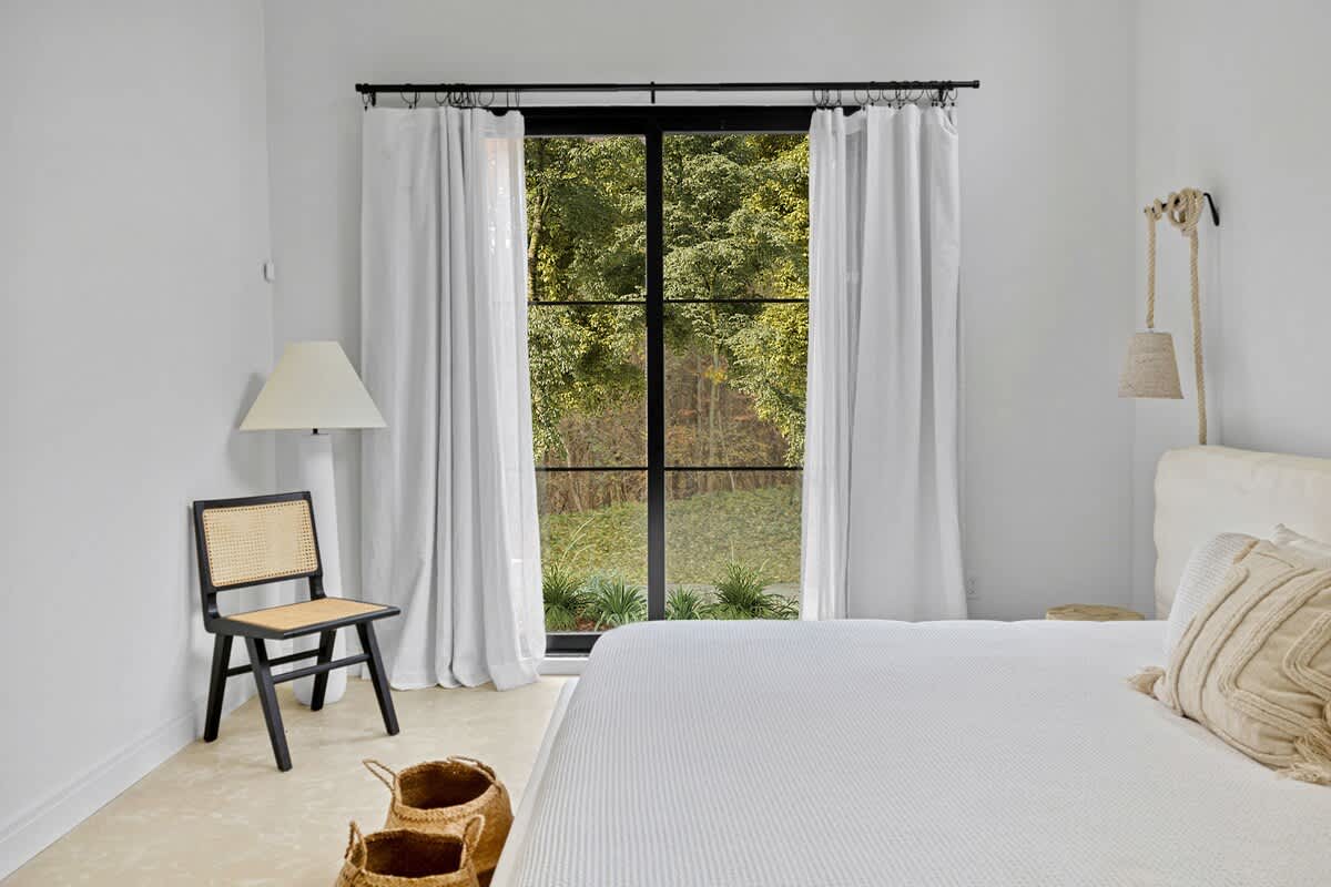 View of a bright bedroom focusing on the large black-framed glass doors with white curtains, offering a lush green outdoor view. A black cane-back chair with a white lamp and woven baskets are visible on the left.