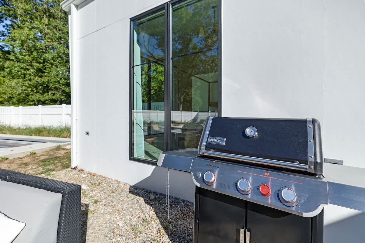 A close-up of a black and stainless steel Weber Genesis BBQ grill stands on a gravel patio next to a white modern building with a large black-framed window. Green foliage and a white fence are visible in the background.