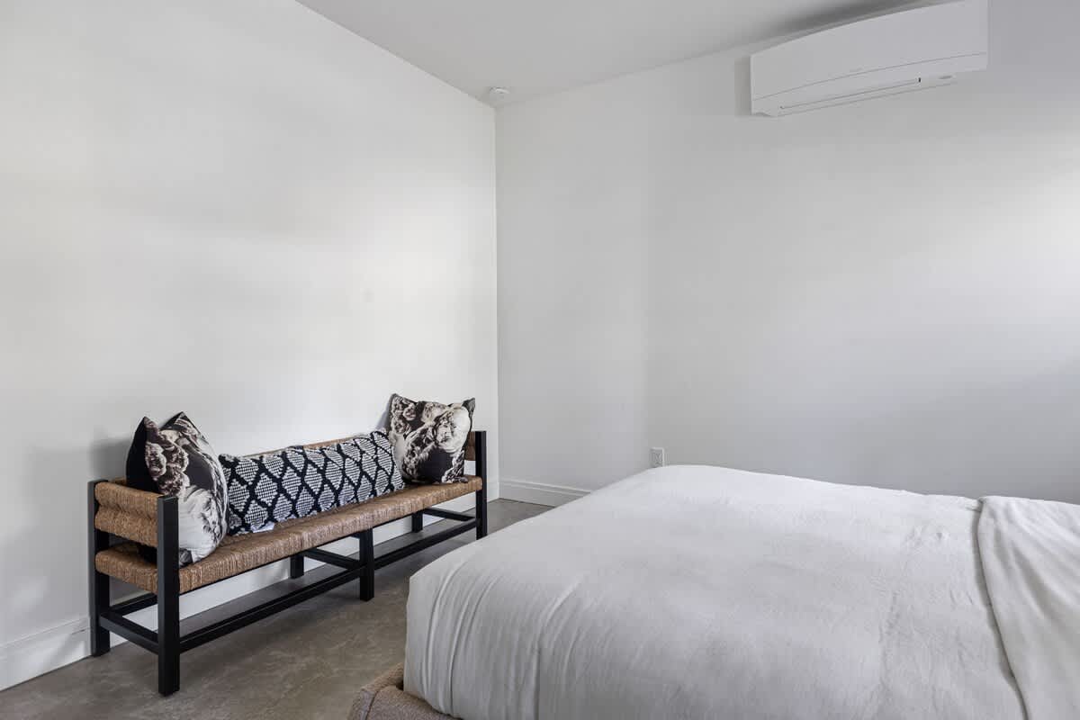 A white-walled bedroom featuring a woven bench with decorative pillows against one wall, and a glimpse of a bed with white bedding, under a wall-mounted air conditioning unit.