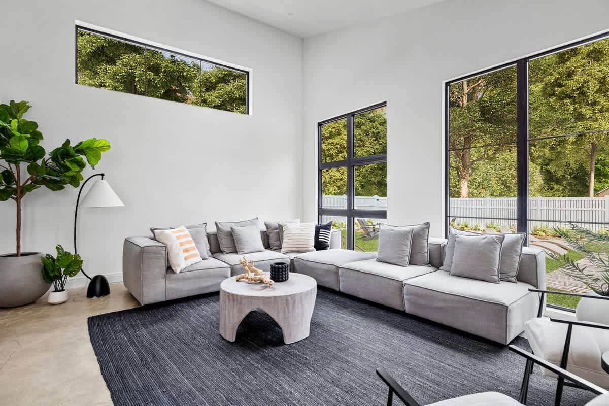 Spacious living room with high ceilings, a large grey modular sectional sofa, a round concrete coffee table, and a dark textured rug, illuminated by tall black-framed windows and a high horizontal window.