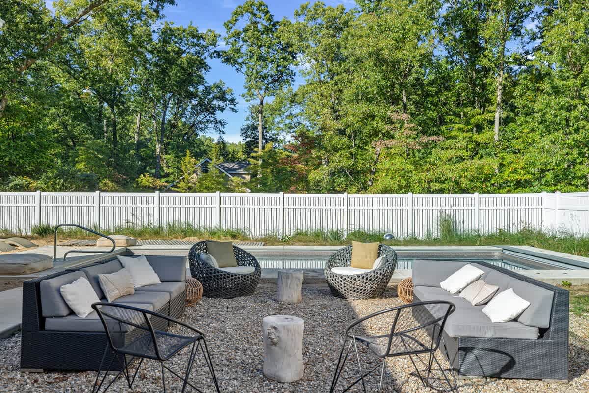 A gravel outdoor seating area features two long modular sofas with grey cushions and two round wicker chairs, centered around three rustic wooden stump tables. A swimming pool and white picket fence are visible in the background.