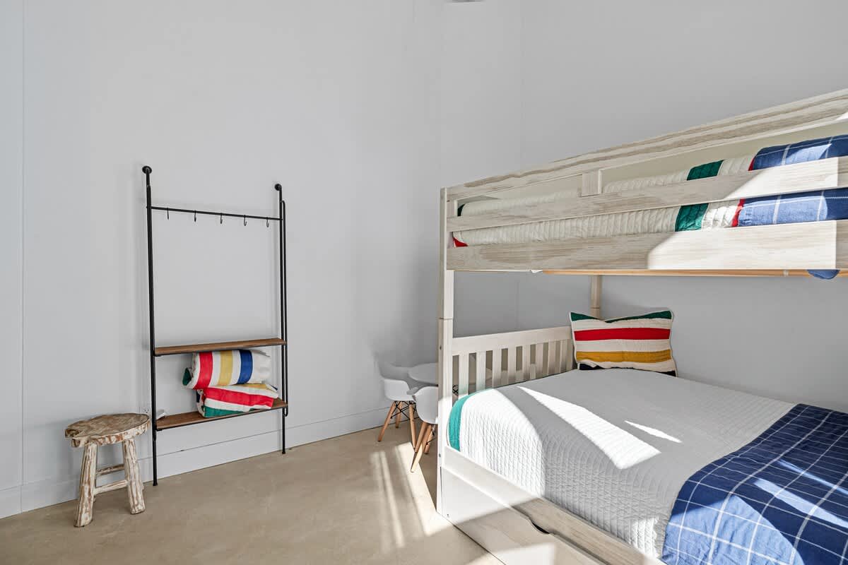 Corner view of a kids' bedroom showing one white wooden bunk bed with colorful bedding, a small white table and chairs, and a black metal shelving unit with rolled towels and pillows.