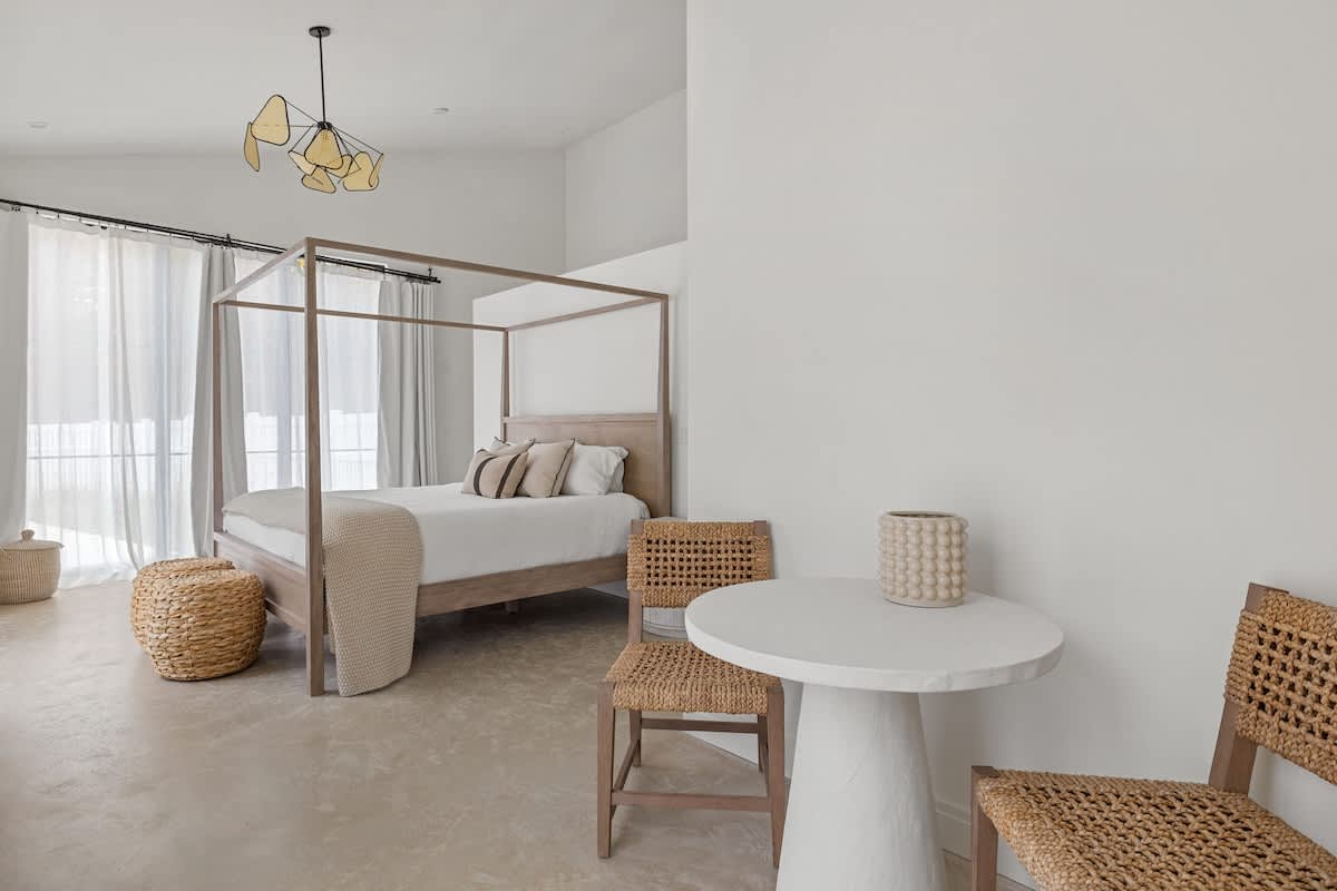 Serene bedroom with a light wood canopy bed dressed in white linens, a small white round table with two woven chairs, and light-colored flooring, creating a bright and airy atmosphere.