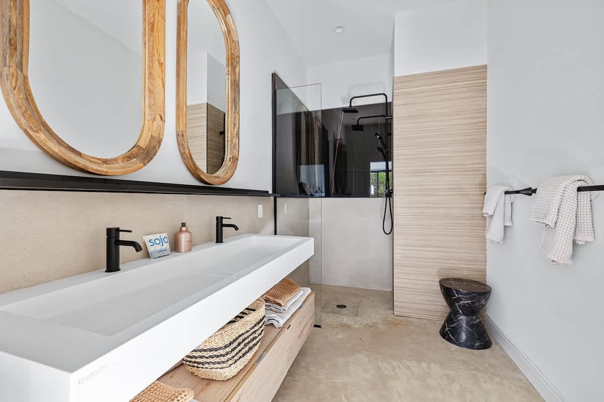 Modern bathroom with a long white trough sink and black faucets, two oval wooden mirrors, and a walk-in shower with a glass partition and dark fixtures, complemented by light wood accents.