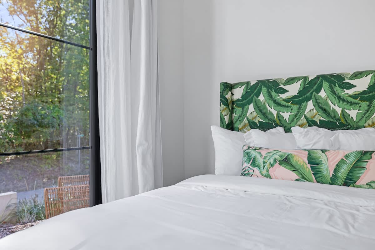 Close-up of a bed in a bright bedroom, showcasing a vibrant green tropical leaf-patterned headboard and a pink and green patterned lumbar pillow, set against a large window with white curtains.