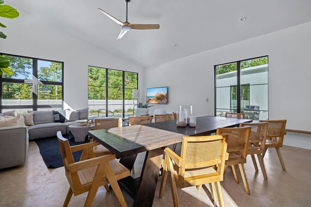 Bright living room with a light gray sectional sofa, a dark area rug, and a large wooden dining table with cane chairs, all illuminated by expansive black-framed windows overlooking lush greenery.