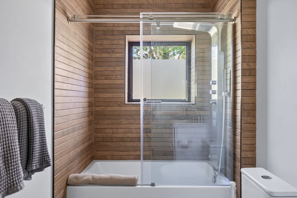 Close-up of the bathtub and shower area in a modern bathroom, featuring horizontal wood-paneled walls, a clear glass shower door, and a frosted window above the tub.