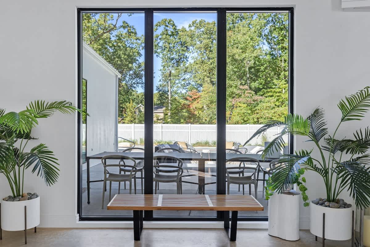 A large floor-to-ceiling window frames an outdoor dining area with a long table and chairs, surrounded by lush green trees and a white fence, creating a seamless indoor-outdoor connection.