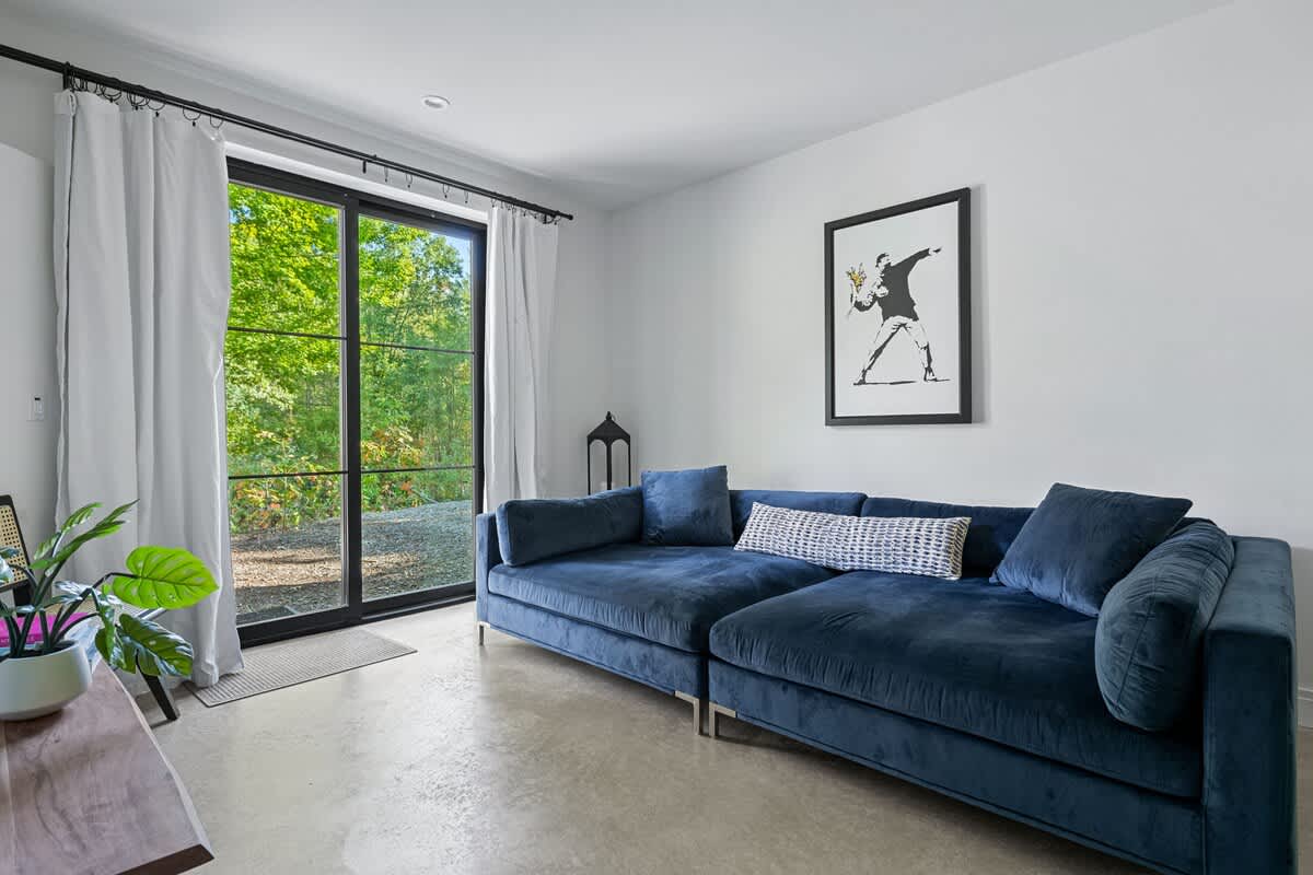 A bright living room with a large blue velvet sectional sofa, a framed Banksy-style artwork on the wall, and a wide glass sliding door offering views of the outdoors.