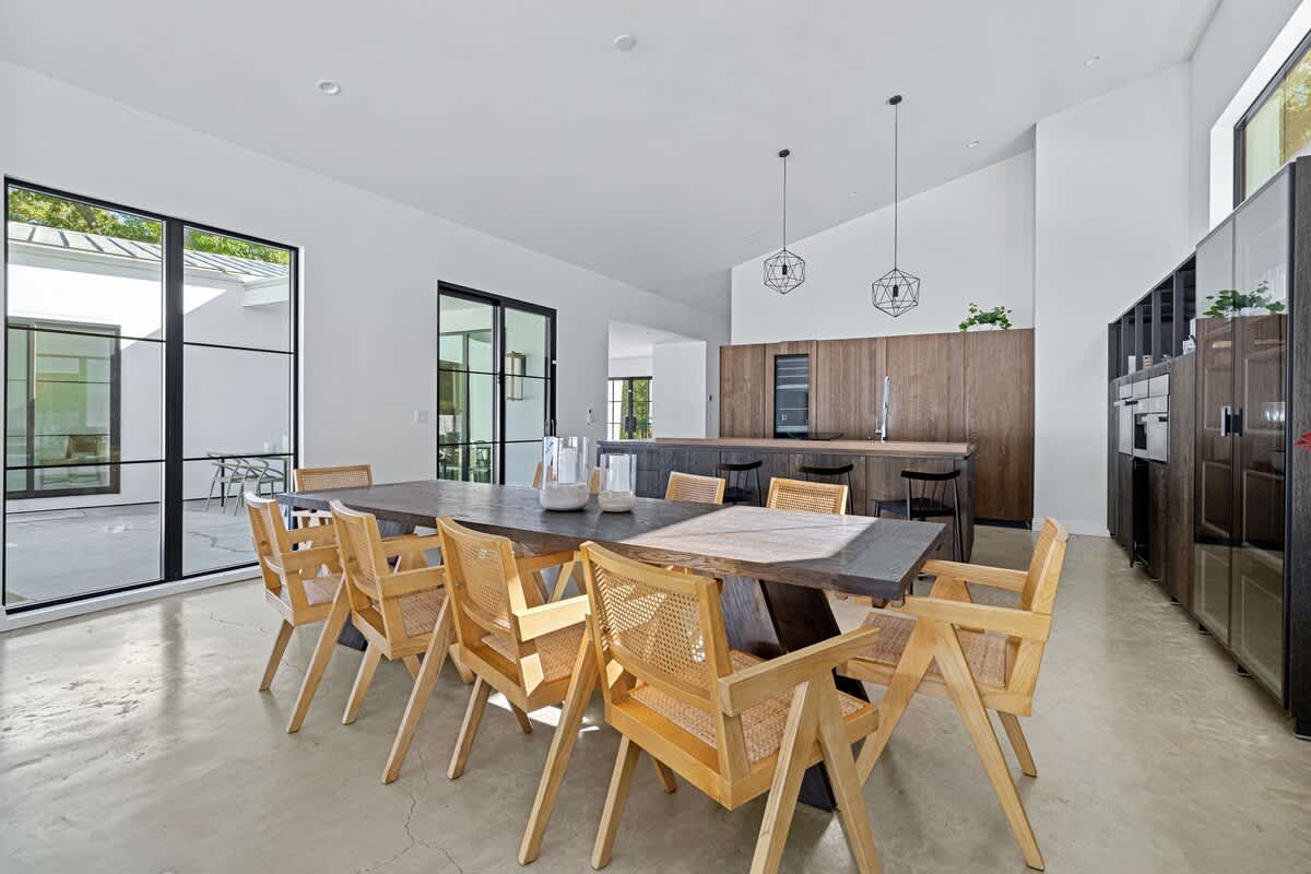 Spacious dining area with a long dark wood table and light wood cane chairs, leading to a modern kitchen island with bar seating and dark wood cabinetry, featuring large black-framed windows.