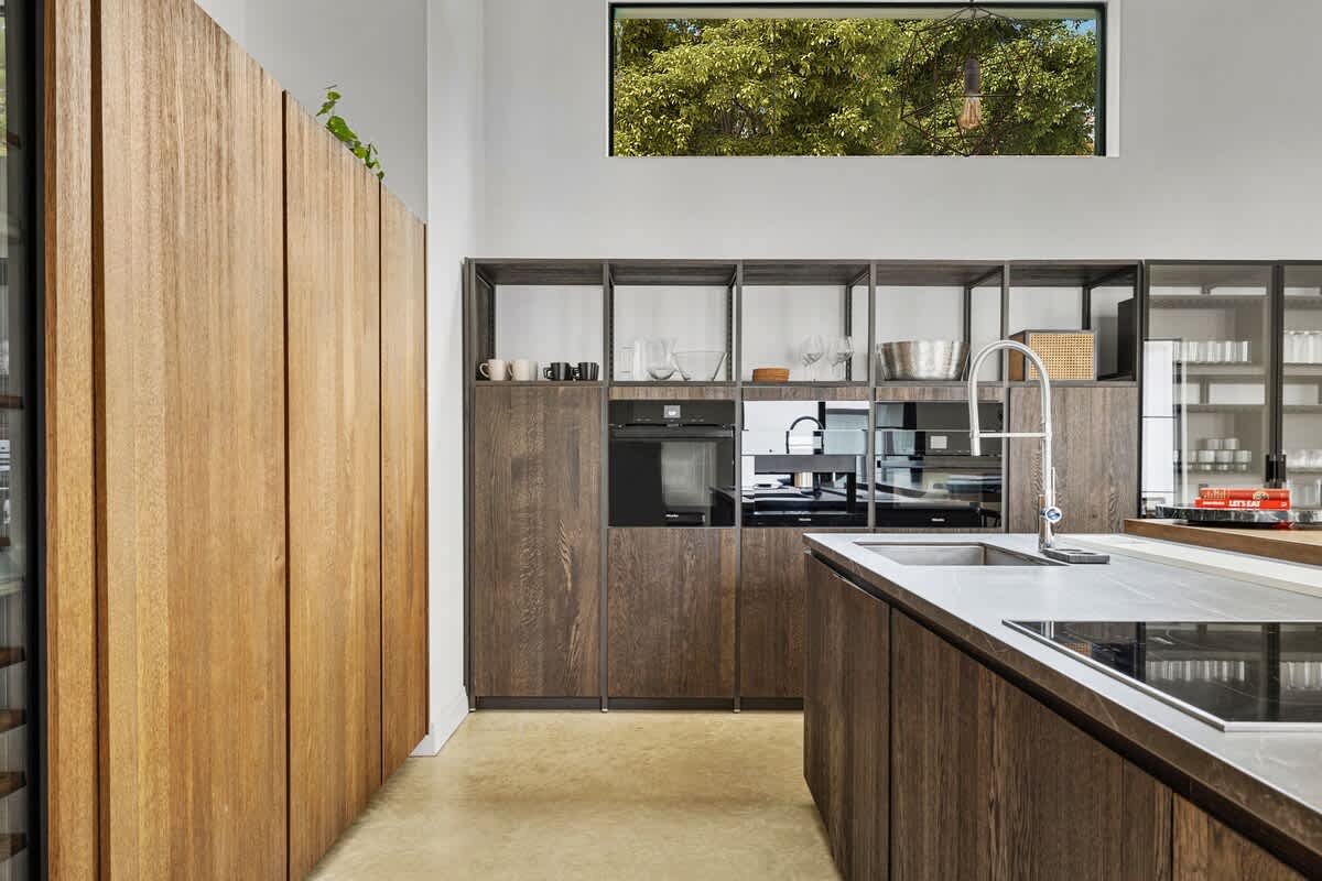 Modern kitchen featuring dark wood cabinetry, integrated appliances, a large island with a sink and cooktop, and a light wood paneled wall, all under a high window.
