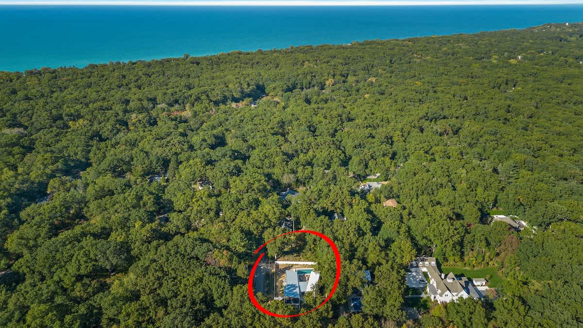 A high aerial view captures the vacation home nestled within a dense green forest, with a red circle highlighting its location. The vast expanse of Lake Michigan is visible in the distance under a clear blue sky.