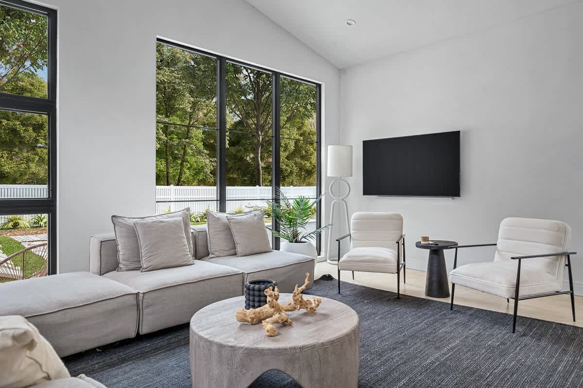 Cozy living area with a grey sectional sofa, two white armchairs, a round concrete coffee table, and a wall-mounted TV, illuminated by large black-framed windows overlooking a fenced yard.