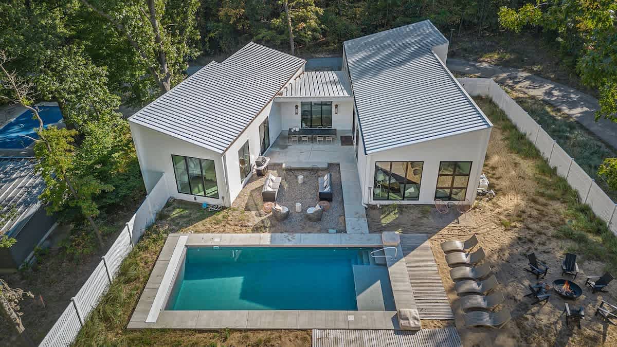 An aerial view showcases the modern vacation home with two distinct white buildings connected by a central patio, all featuring angled metal roofs. A rectangular swimming pool, fire pit area, and surrounding wooded landscape are clearly visible.