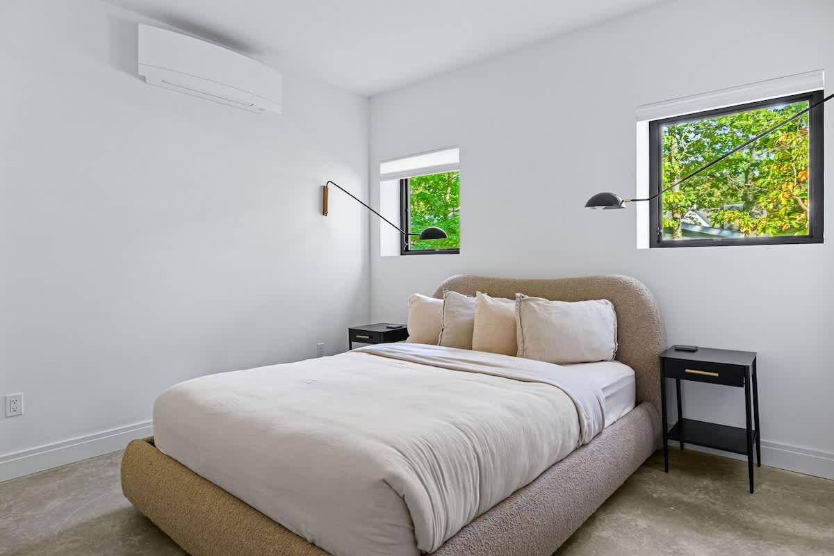 A bright bedroom with white walls, a beige upholstered bed, two black nightstands, and modern wall sconces flanking the bed, with two windows offering views of lush greenery.
