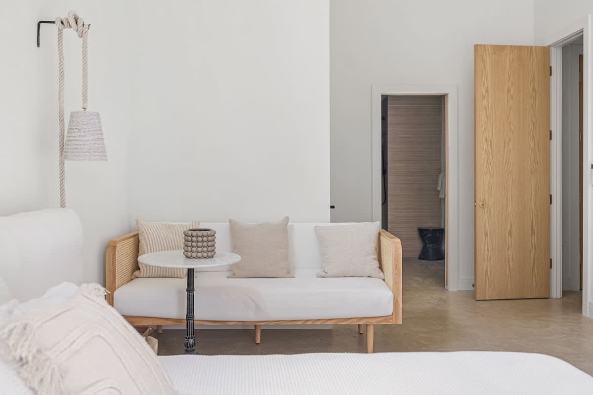 Cozy corner of a bedroom featuring a light wood daybed with neutral cushions, a small marble-top side table, and a unique rope-style wall lamp, with a light wood door leading to a bathroom.