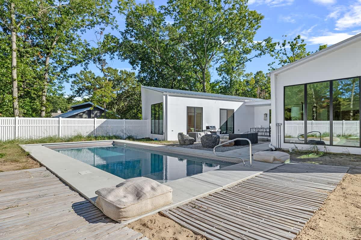 A rectangular swimming pool with blue water is surrounded by light concrete decking and wooden planks, set in a sandy yard. The modern white house with black-framed windows is visible in the background, along with a white picket fence and lush trees.