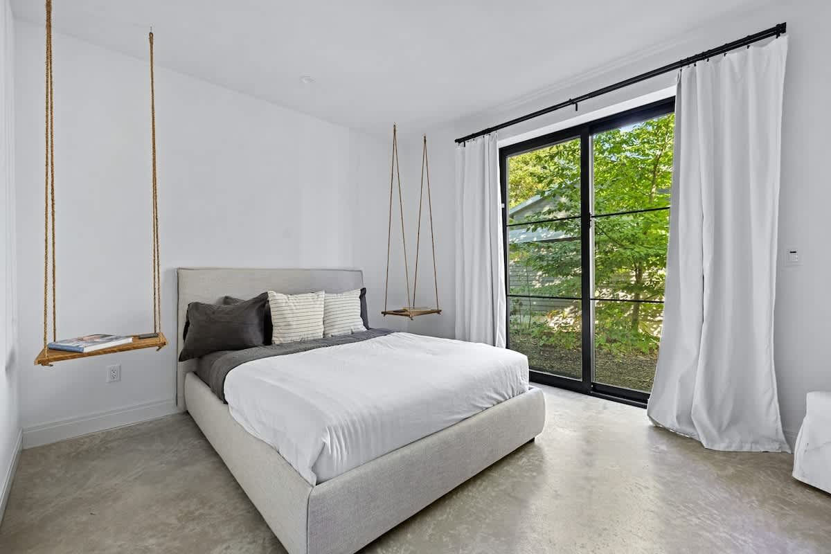 A modern bedroom with a gray upholstered bed, white bedding, and unique hanging wooden shelf nightstands, set against a large glass sliding door overlooking lush greenery.