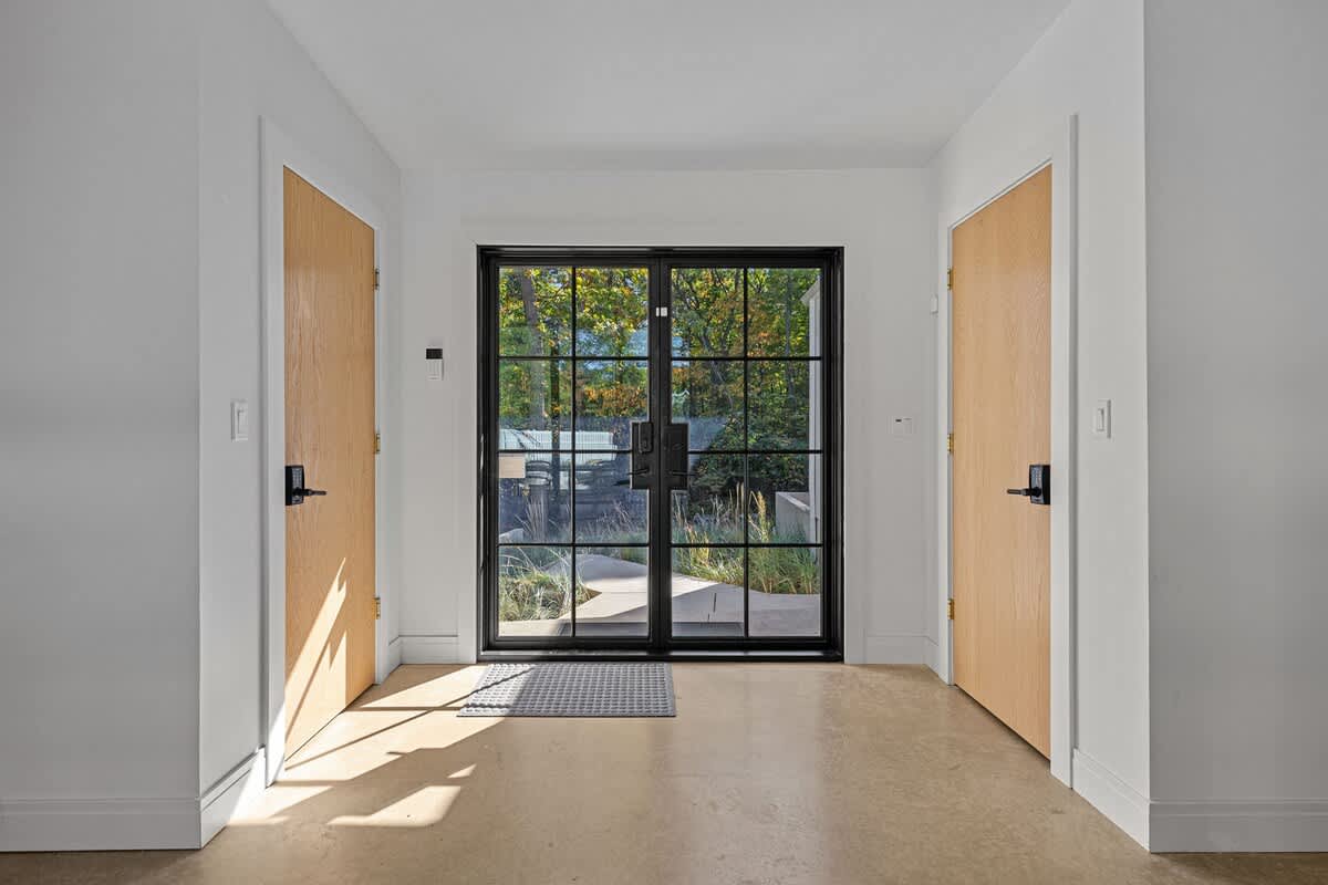 Bright entryway with a large black-framed glass double door offering views to the outside, flanked by two light wood doors, and featuring light-colored flooring and white walls.