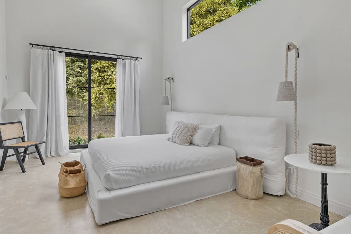 Serene bedroom with a white upholstered bed, white bedding, and large black-framed glass doors offering a view of the surrounding trees. Rope-hung sconces flank the bed, and a black chair with a cane back sits in the corner.