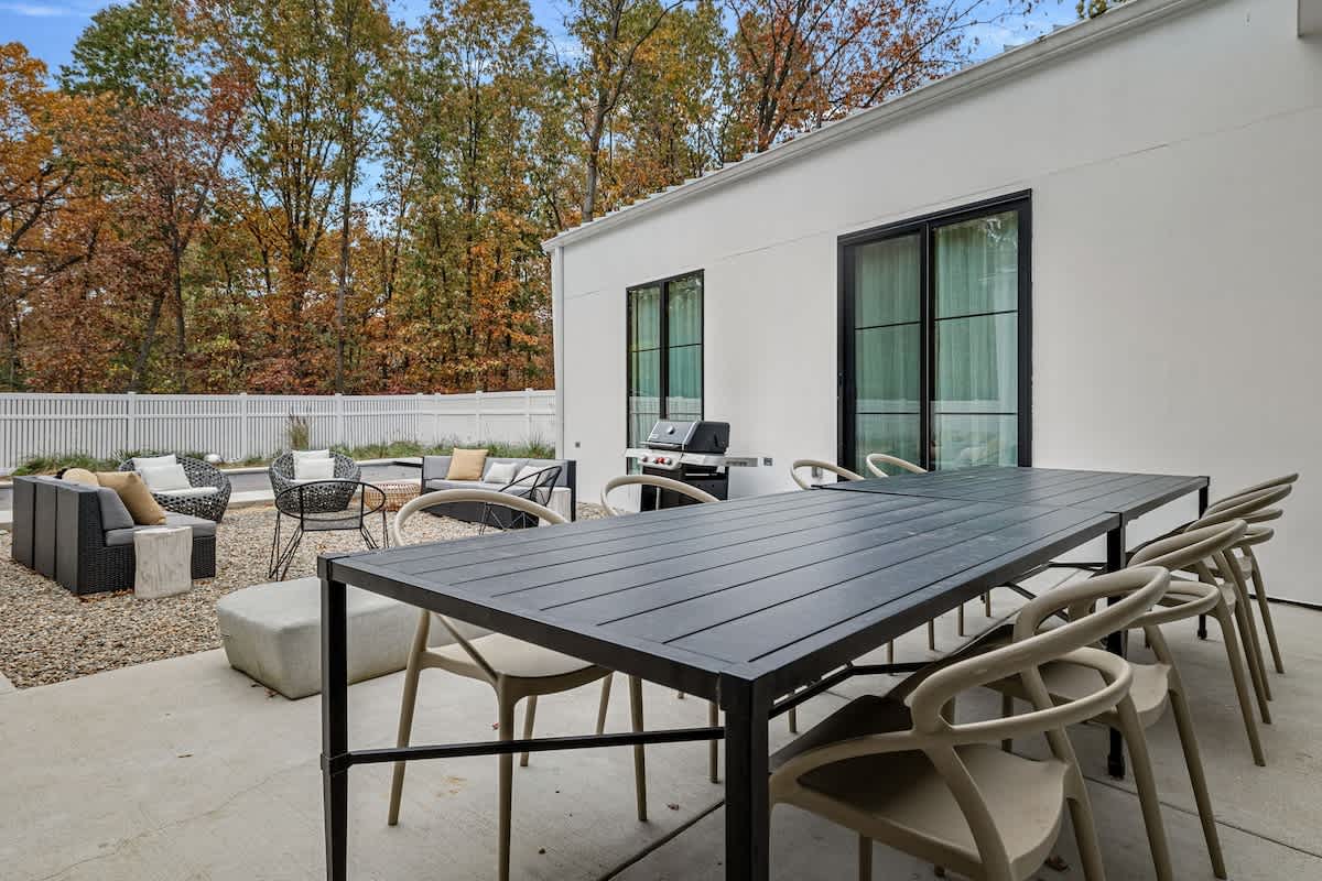 Expansive outdoor patio featuring a long black dining table with multiple chairs and a separate lounge area with a sectional sofa and armchairs, all set on concrete and gravel, surrounded by a white fence and trees.