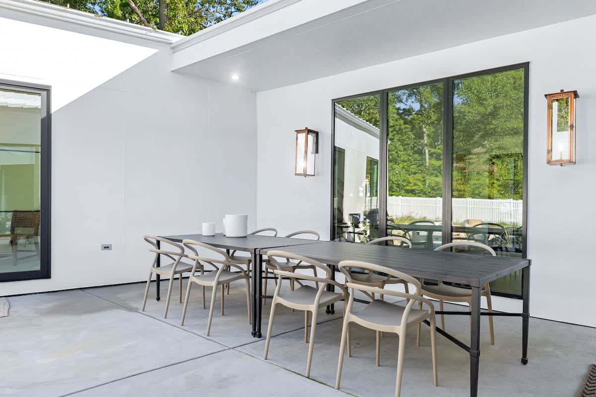 A covered outdoor dining area features a long dark table with multiple light-colored chairs, set against a white wall with large black-framed windows. Two modern wall sconces illuminate the space, which has a smooth concrete floor.