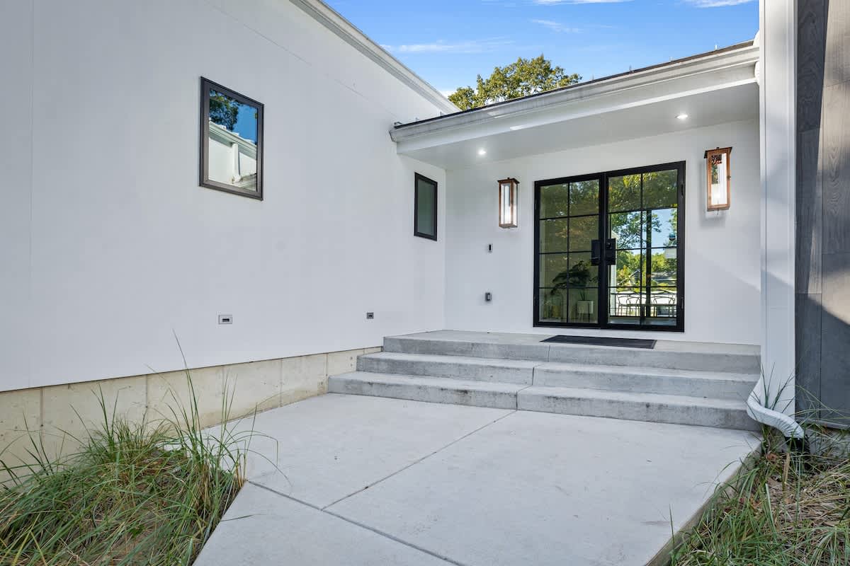 Concrete patio leading to a modern entrance with black-framed double doors and sconce lighting, flanked by white stucco walls and small windows, with grass and landscaping in the foreground.