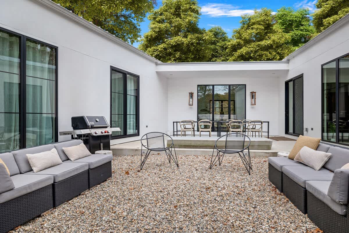 A modern outdoor courtyard features white walls, large black-framed windows, and a gravel ground. It includes two long modular sofas, two round metal chairs, and a BBQ grill, with a dining area visible in the background.
