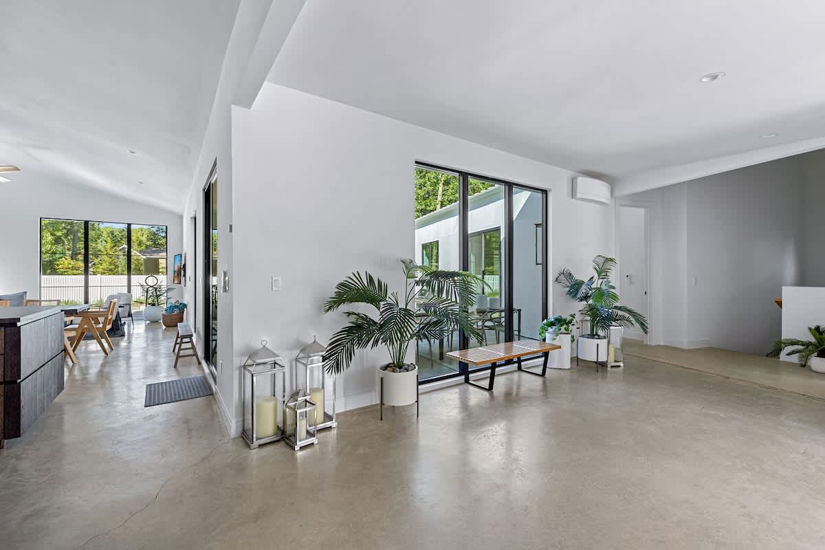 An expansive open-concept living area with polished concrete floors, featuring large windows overlooking greenery, decorative plants in white planters, and a hallway leading to other parts of the house.