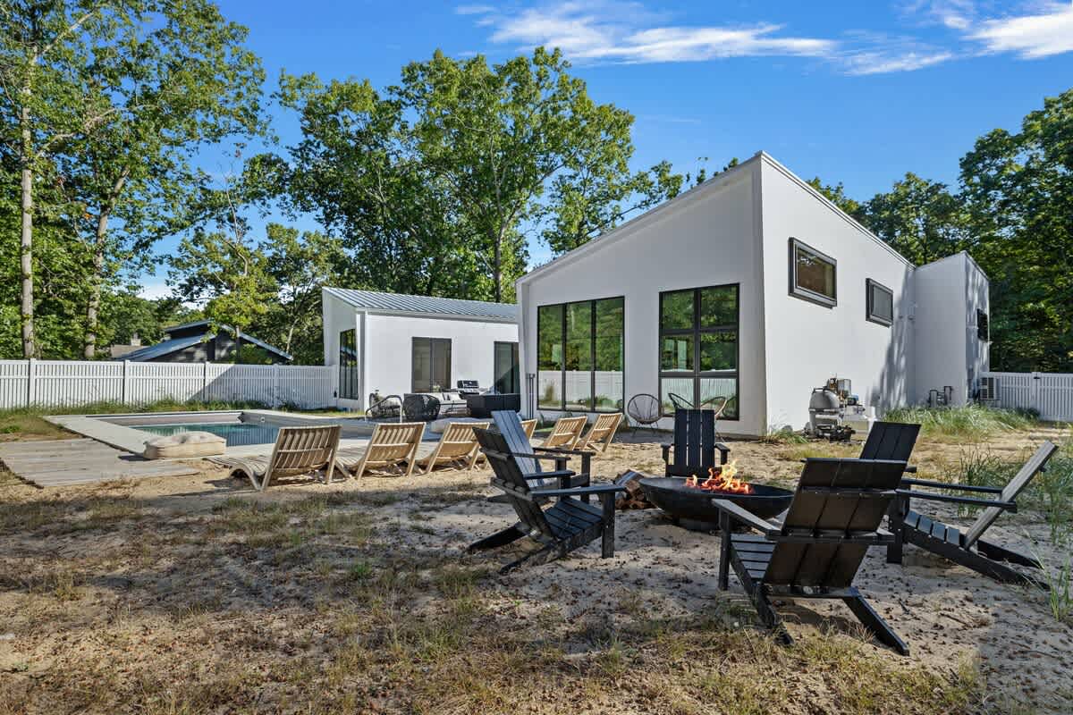 An outdoor fire pit area features a black metal fire pit surrounded by dark Adirondack-style chairs on a sandy ground. The modern white house with black-framed windows and a swimming pool are visible in the background, surrounded by trees.