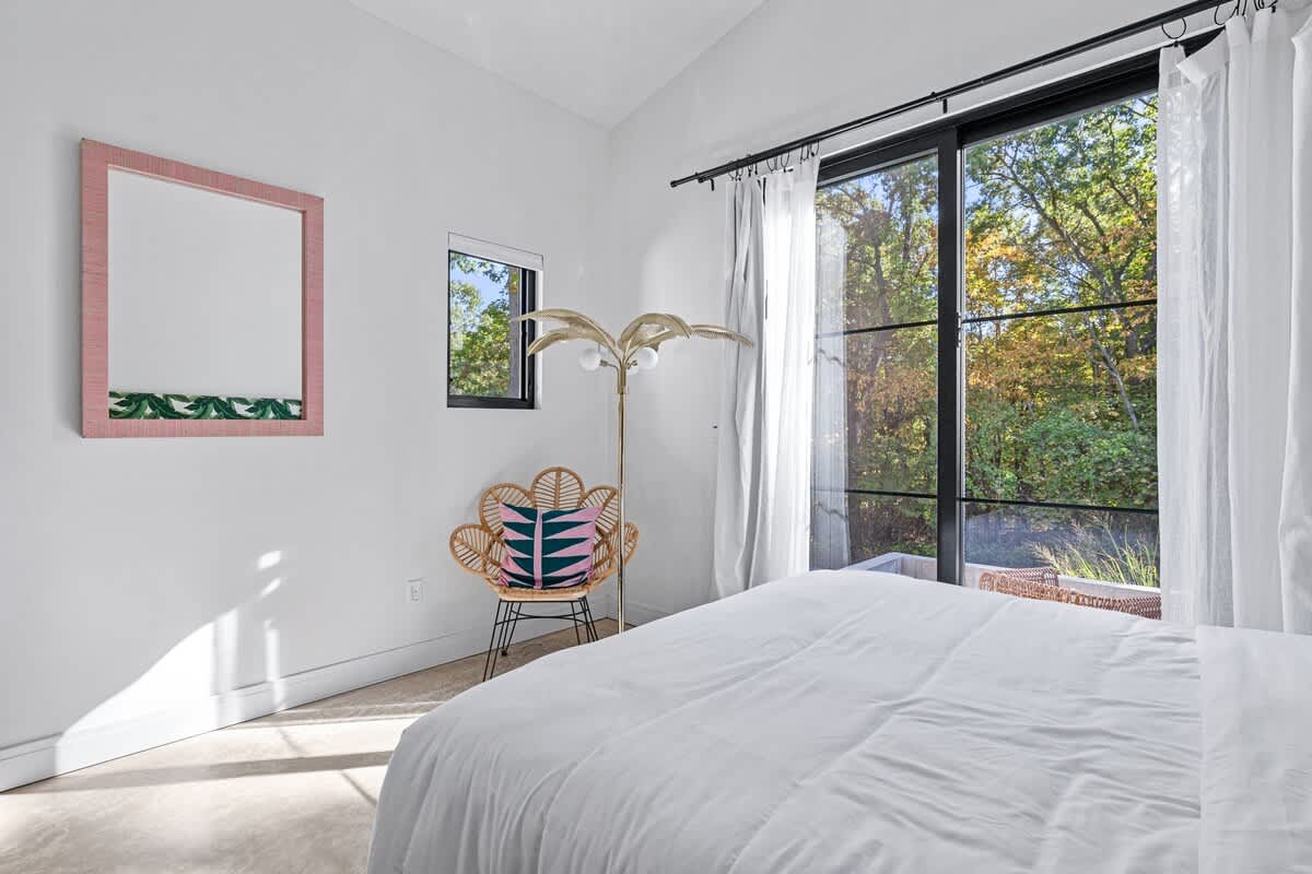 Bright bedroom with white walls, a large bed with white bedding, and a black-framed glass door leading to an outdoor area with lush greenery. A pink-framed mirror and a rattan chair with a colorful pillow add a touch of tropical style.
