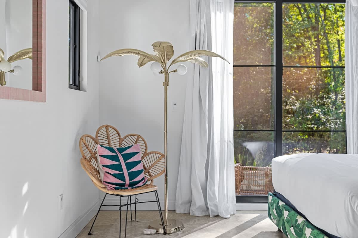 Close-up view of the tropical-themed corner in a bright bedroom, featuring a rattan flower-shaped chair with a pink and teal geometric pillow, a golden palm tree floor lamp, and a large glass door revealing lush outdoor foliage.